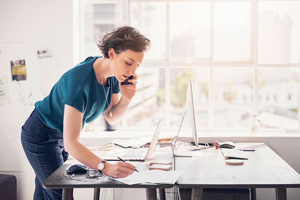 Frau telefoniert am Arbeitsplatz und notiert sich nebenbei etwas. Am Tisch stehen ein Laptop und ein PC sowie weitere Arbeitsutensilien.