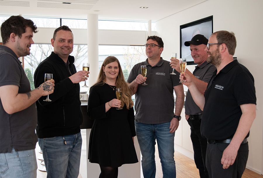 Fünf Männer und eine Frau stehen mit einem Glas Sekt in der Hand lachend da