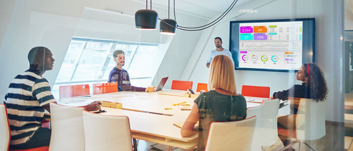 Fünf Personen unterhalten sich in einem Besprechungsraum. An der Wand hängt ein Fernseher, der die Kennzahlen aus der Dashboard-Software zeigt.